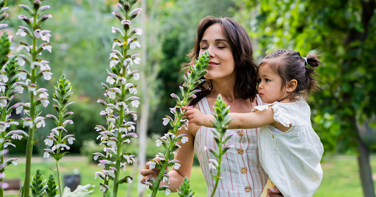 Side view of relaxed mother with active child in hands smelling flowers with closed eyes and enjoying fragrance in green park